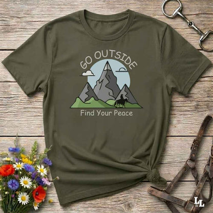 An olive green graphic t-shirt laid flat on a rustic wooden surface, styled with a small bouquet of flowers, a metal horse bit, and leather straps. The t-shirt features a graphic of gray mountains, green hills, and a light blue sun with the text 'GO OUTSIDE Find Your Peace'.
