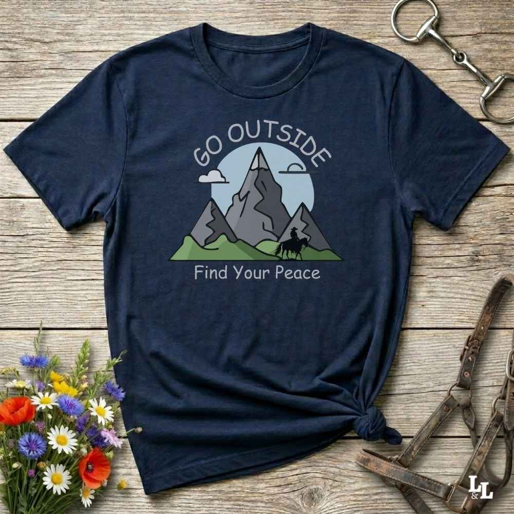 A navy blue graphic t-shirt laid flat on a rustic wooden surface, styled with a small bouquet of wildflowers on the left and leather tack on the right. The t-shirt features a graphic of gray mountains, green hills, a light blue sun, and the text 'GO OUTSIDE Find Your Peace'.