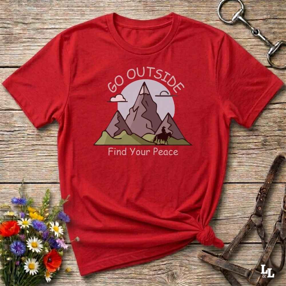 A red short-sleeve graphic T-shirt laid flat on a rustic wooden surface, with a mountain illustration and the words 'GO OUTSIDE Find Your Peace' printed on the front. The shirt is styled with flowers, a metal key ring, and leather straps around it.