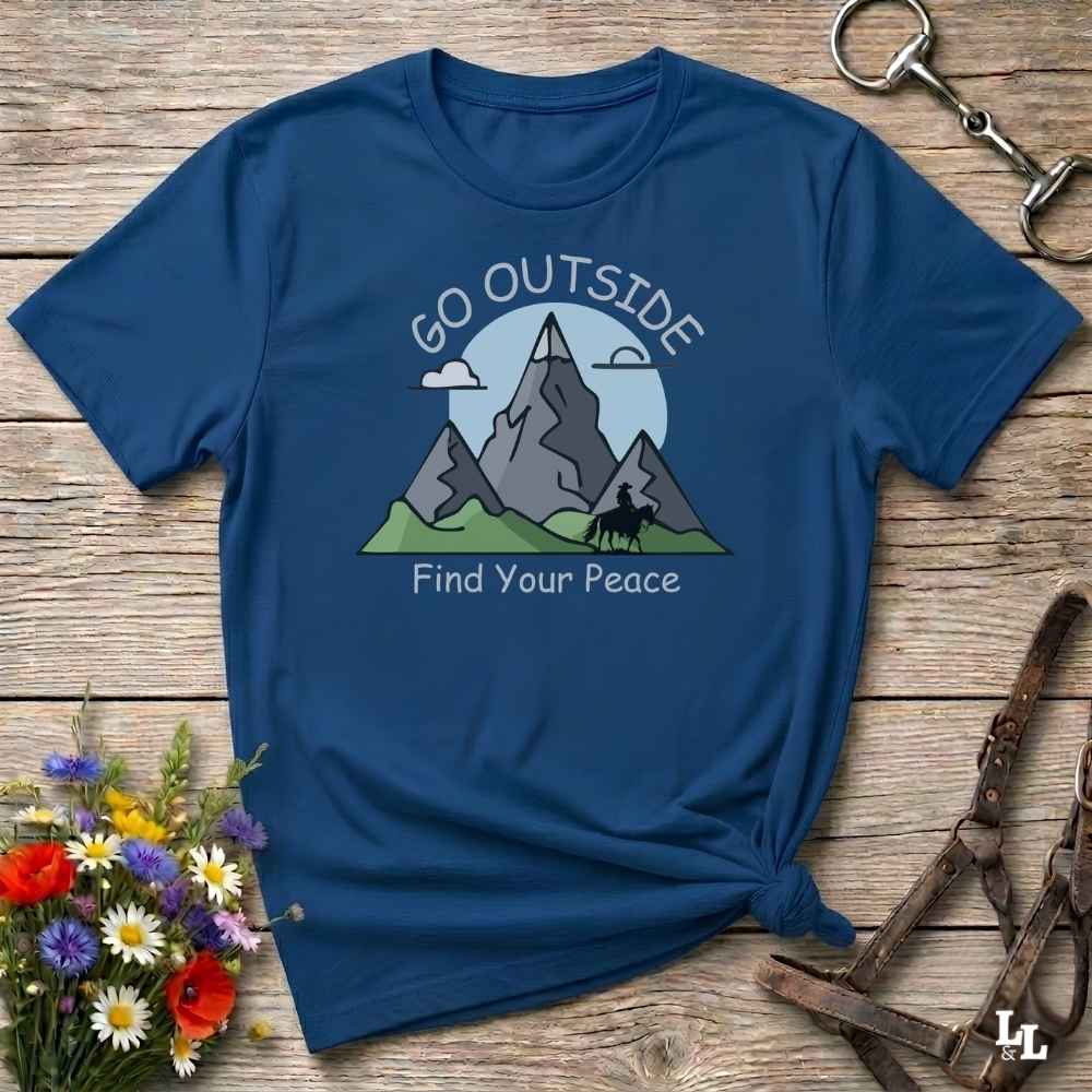 A navy blue graphic t-shirt laid flat on a rustic wooden surface, styled with wildflowers, a horse bit, and a leather halter. The t-shirt features a mountain graphic with the text 'GO OUTSIDE Find Your Peace'.