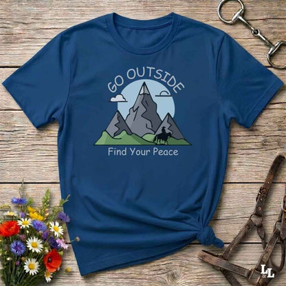 A navy blue graphic t-shirt laid flat on a rustic wooden surface, styled with wildflowers, a horse bit, and a leather halter. The t-shirt features a mountain graphic with the text 'GO OUTSIDE Find Your Peace'.