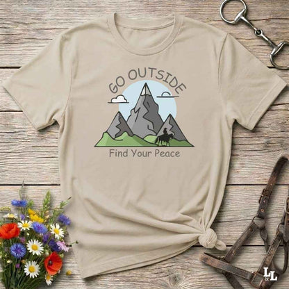 A beige graphic t-shirt with a mountain design and the text 'GO OUTSIDE Find Your Peace', styled in a flat-lay on a rustic wooden background with flowers and leather accessories.