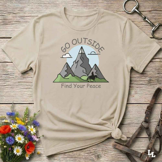 A beige graphic t-shirt with a mountain design and the text 'GO OUTSIDE Find Your Peace', styled in a flat-lay on a rustic wooden background with flowers and leather accessories.