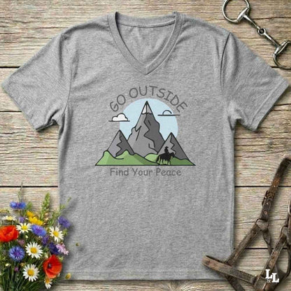 A heather gray V-neck t-shirt with a graphic of mountains, a horse silhouette, and the text 'GO OUTSIDE Find Your Peace', displayed flat on a rustic wooden background with equestrian and floral props.