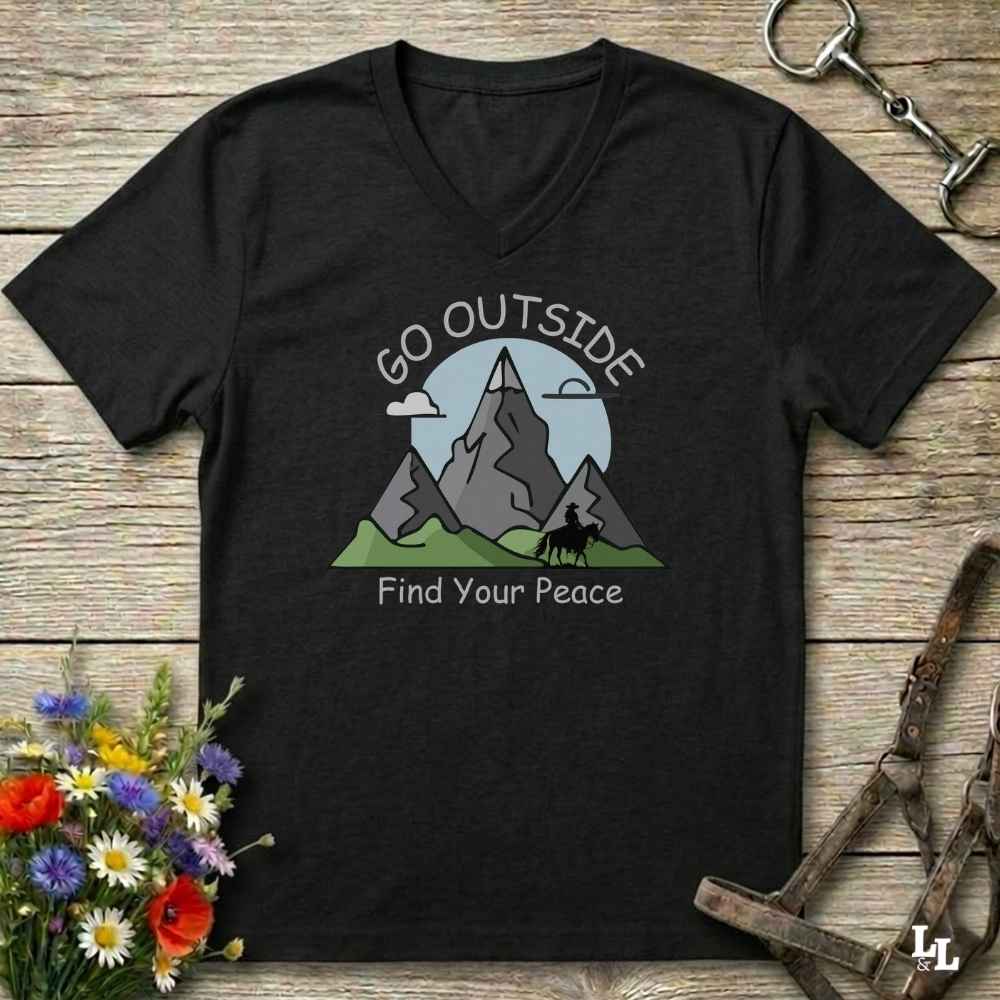 A black V-neck t-shirt with a mountain graphic and the text 'GO OUTSIDE Find Your Peace' printed on the chest, displayed on a rustic wooden background with decorative flowers and leather gear.