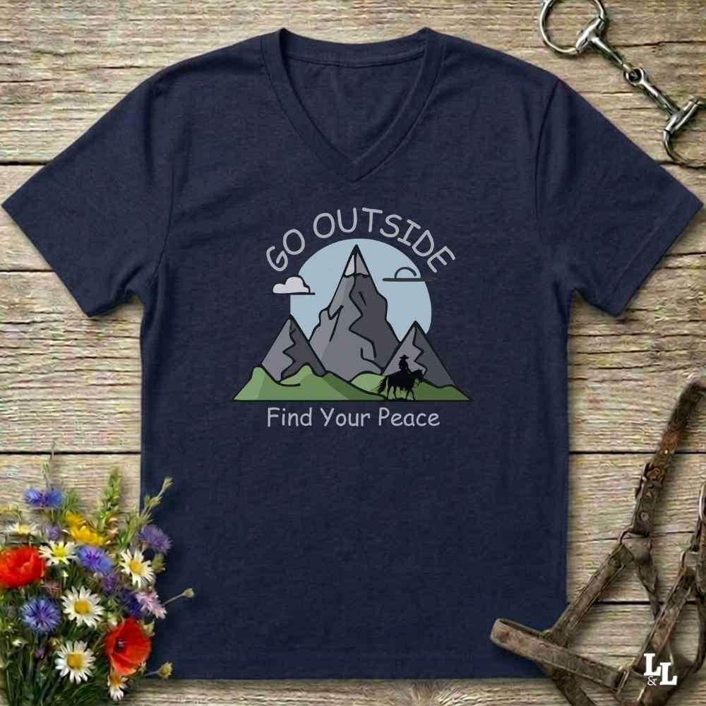 A navy blue V-neck t-shirt with a graphic of mountains, a horse rider, and the text 'GO OUTSIDE Find Your Peace', laid flat on a rustic wooden background with decorative flowers and tack.