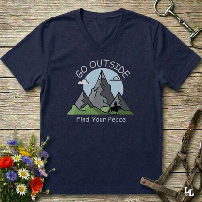 A navy blue V-neck t-shirt with a graphic of mountains, a horse rider, and the text 'GO OUTSIDE Find Your Peace', laid flat on a rustic wooden background with decorative flowers and tack.