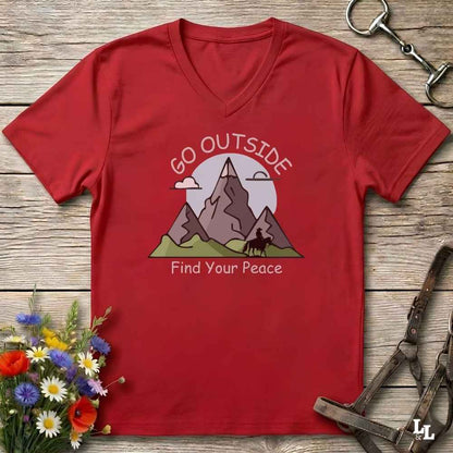 A red V-neck t-shirt laid flat on a rustic wooden surface, featuring a graphic of mountains, a horse, and the text 'GO OUTSIDE Find Your Peace'. The shirt is styled with equestrian tack and a small bouquet of wildflowers.