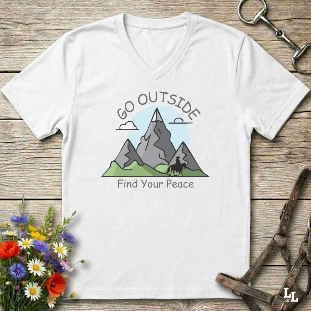 A white V-neck t-shirt laid flat on a rustic wooden surface, featuring a graphic of gray mountains, green hills, and a horse silhouette with the text 'GO OUTSIDE Find Your Peace'. The shirt is styled with equestrian hardware and flowers.