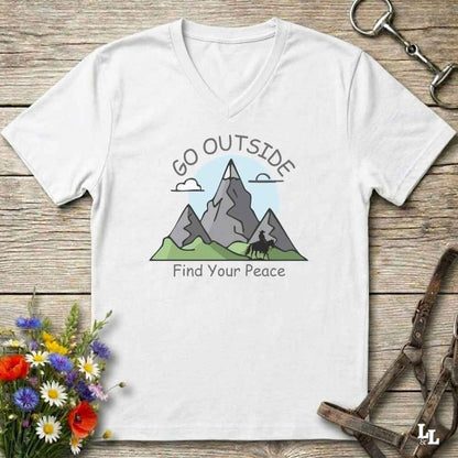 A white V-neck t-shirt laid flat on a rustic wooden surface, featuring a graphic of gray mountains, green hills, and a horse silhouette with the text 'GO OUTSIDE Find Your Peace'. The shirt is styled with equestrian hardware and flowers.