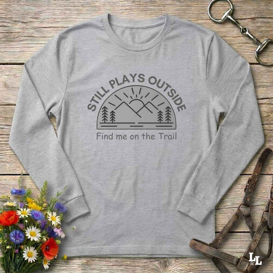 A light heather gray long-sleeve t-shirt with a dark gray graphic print reading 'STILL PLAYS OUTSIDE' and 'Find me on the Trail' with a mountain and tree silhouette. The shirt is displayed flat on a rustic wooden background with decorative props including a bouquet of flowers, a metal tack item, and a brown leather strap.