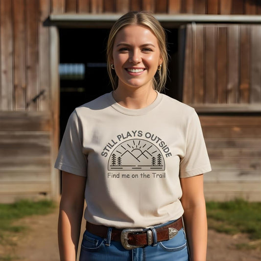 Woman wearing a t-shirt with 'Still Plays Outside, Find me on the Trail' design in front of a wooden building. Still Plays Outside T-Shirt for Horse Lovers, Trail Riding Tee for Horse Riders