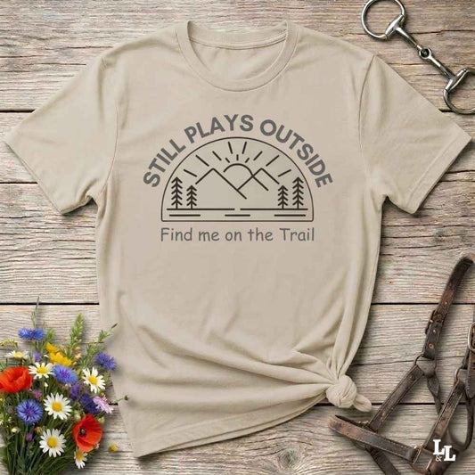 A beige graphic t-shirt with a mountain and sun design, laid flat on a rustic wooden surface with a small knot at the bottom. The text reads 'STILL PLAYS OUTSIDE' and 'Find me on the Trail'.