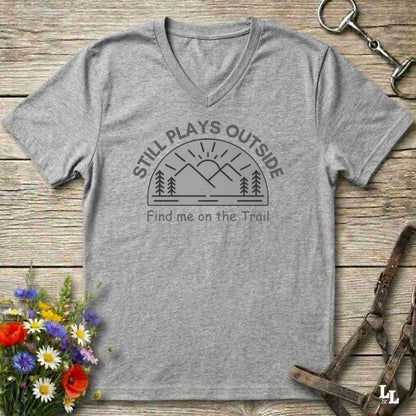 A heather gray V-neck t-shirt with a dark gray graphic reading 'STILL PLAYS OUTSIDE' and 'Find me on the Trail' above a mountain and sun illustration. The shirt is laid flat on a rustic wooden surface with equestrian tack and a small flower arrangement around it.