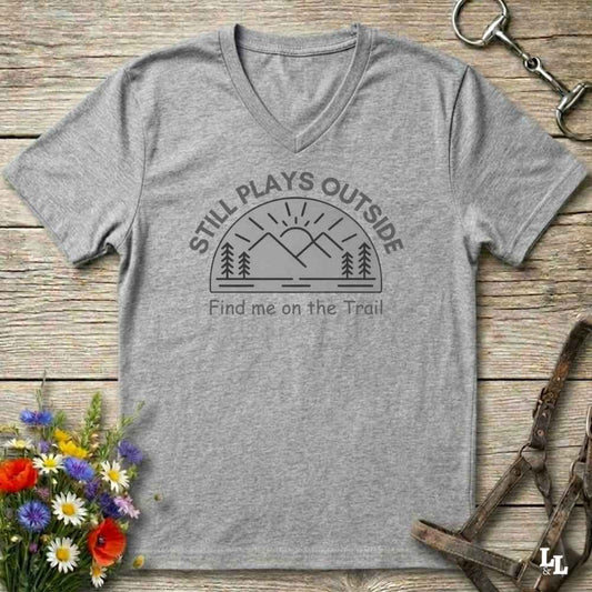 A heather gray V-neck t-shirt with a dark gray graphic reading 'STILL PLAYS OUTSIDE' and 'Find me on the Trail' above a mountain and sun illustration. The shirt is laid flat on a rustic wooden surface with equestrian tack and a small flower arrangement around it.
