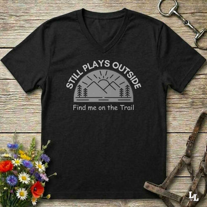 A black V-neck t-shirt with a gray graphic of mountains, trees, and a sun, with the text 'STILL PLAYS OUTSIDE' and 'Find me on the Trail', laid flat on a rustic wood background with flowers and horse tack props.