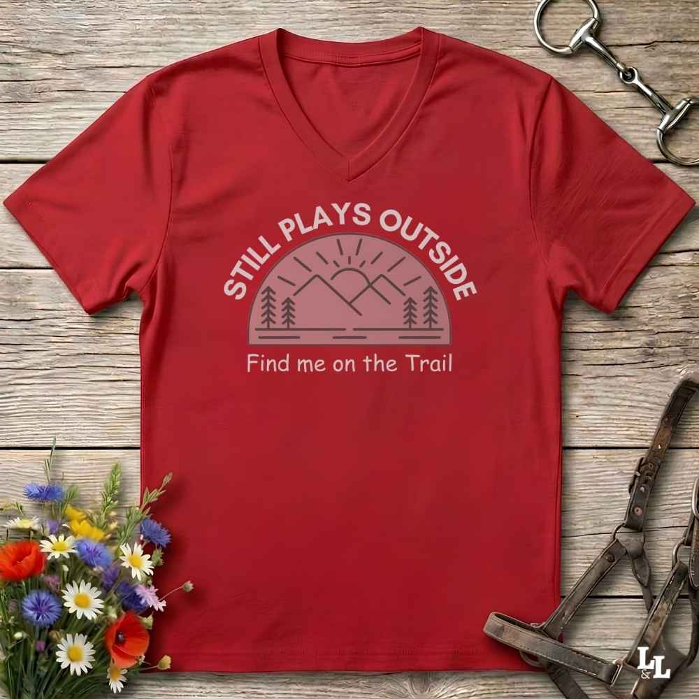 A red V-neck t-shirt laid flat on a rustic wooden surface, featuring a dusty pink graphic of mountains and trees with the text 'STILL PLAYS OUTSIDE' and 'Find me on the Trail'. The shirt is styled with equestrian tack in the top right and a small bouquet of wildflowers in the bottom left.