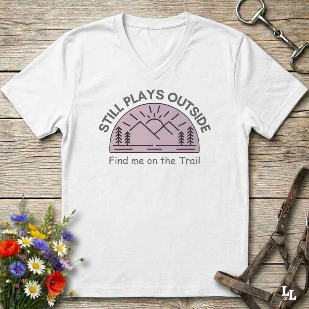 A white v-neck t-shirt laid flat on a rustic wooden surface, featuring a graphic of a mountain and trees with the text 'STILL PLAYS OUTSIDE' and 'Find me on the Trail'. The shirt is styled with a bouquet of wildflowers, a horse bit, and a leather strap in the corners.