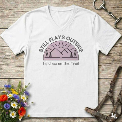 A white v-neck t-shirt laid flat on a rustic wooden surface, featuring a graphic of a mountain and trees with the text 'STILL PLAYS OUTSIDE' and 'Find me on the Trail'. The shirt is styled with a bouquet of wildflowers, a horse bit, and a leather strap in the corners.