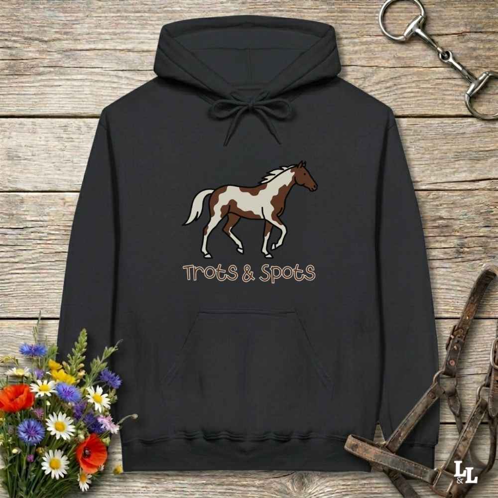 A black pullover hoodie laid flat on a rustic wood surface, featuring a brown and white paint horse graphic with the words 'Trots & Spots' underneath. The hoodie is styled with floral accents and horse tack props.