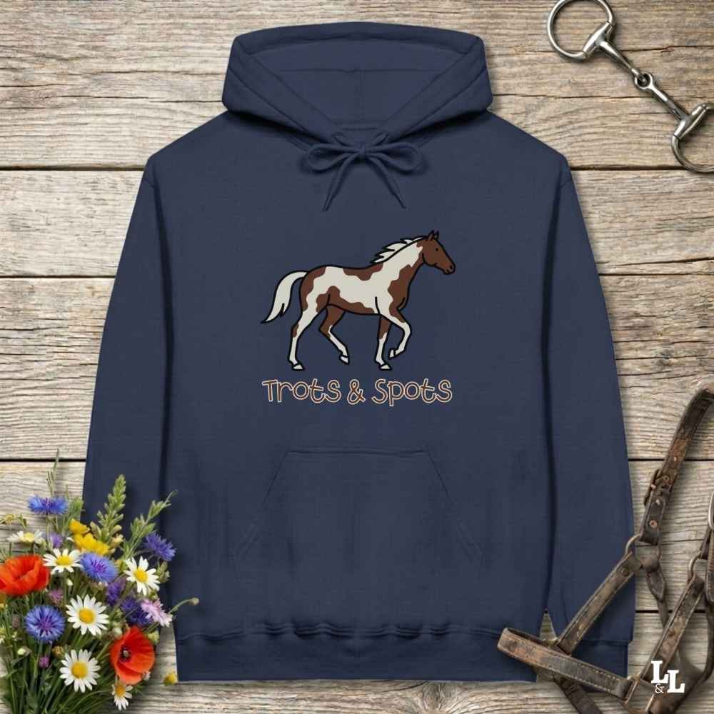 A navy blue hoodie with a brown and white paint horse graphic and the text 'Trots & Spots' printed on the chest, displayed on a rustic wooden background with decorative flowers and horse tack.