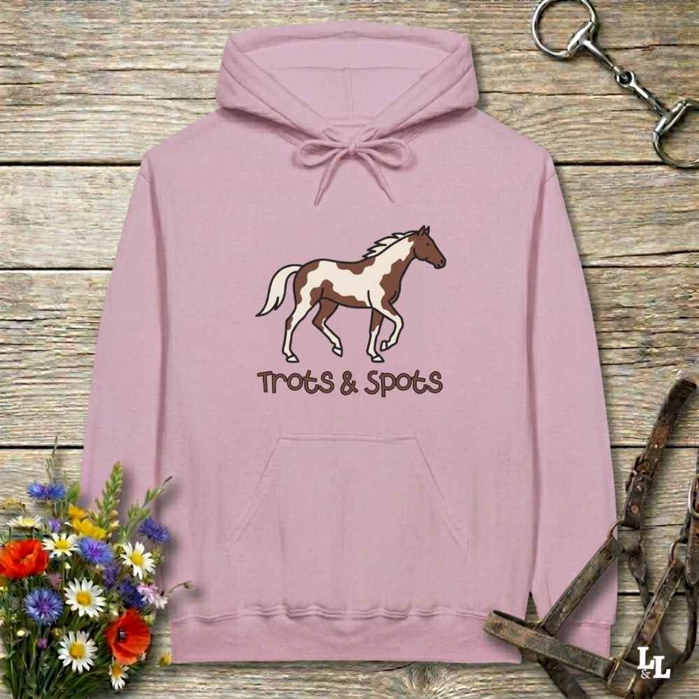 A dusty pink pullover hoodie laid flat on a rustic wooden surface, featuring a brown and white pinto horse graphic with the text 'Trots & Spots' underneath. The hoodie is styled with equestrian-themed props including a halter, reins, and a bouquet of wildflowers.