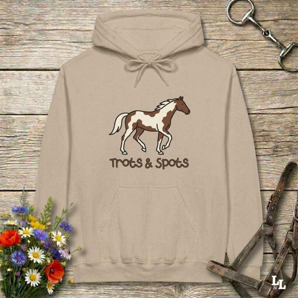 A beige pullover hoodie laid flat on a rustic wood surface, featuring a brown and white pinto horse illustration with the words 'Trots & Spots' underneath. The hoodie is styled with equestrian accessories and flowers around it.