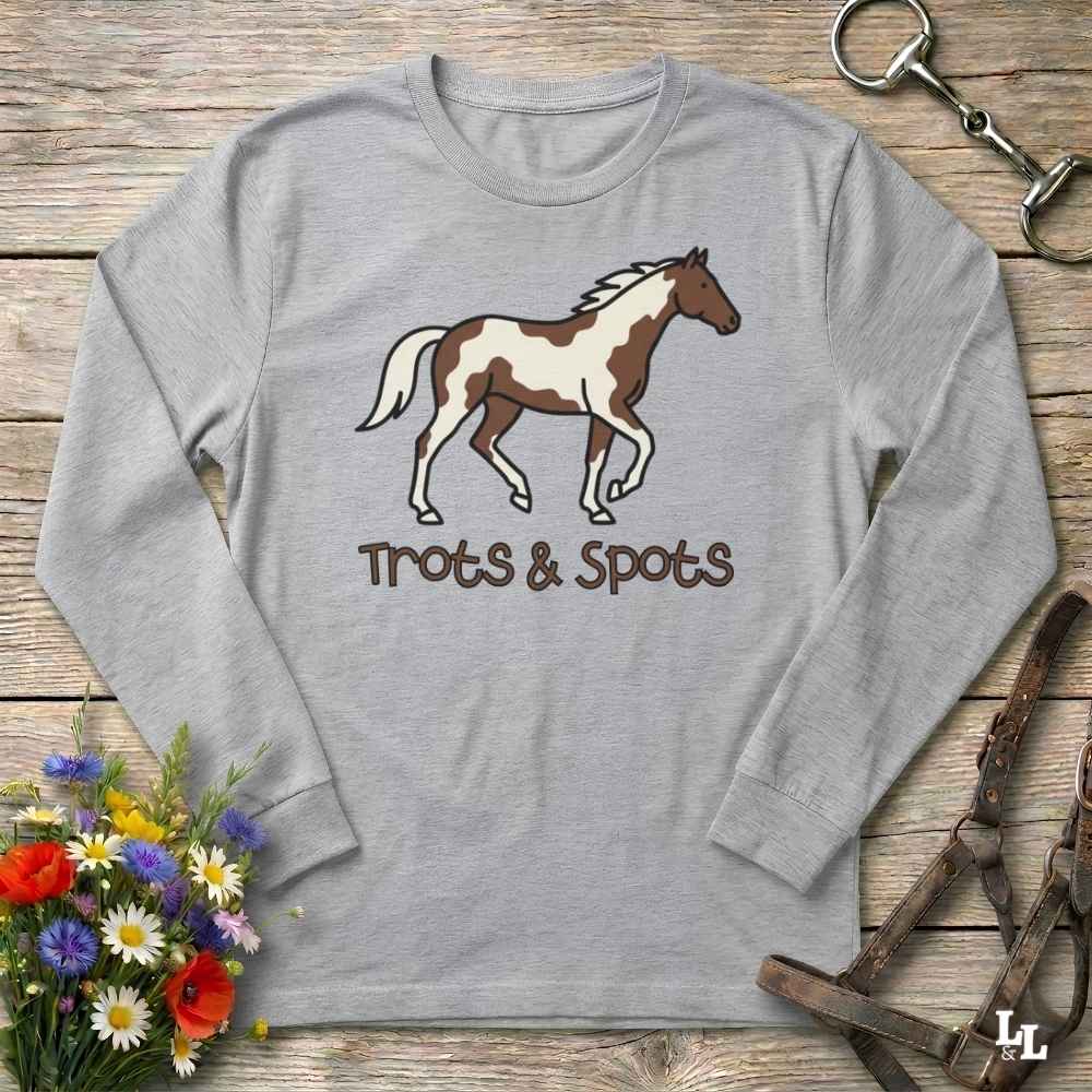 A light heather gray long-sleeve t-shirt with a brown and white paint horse graphic and the text 'Trots & Spots' printed on the front, displayed on a rustic wooden background with equestrian and floral props.