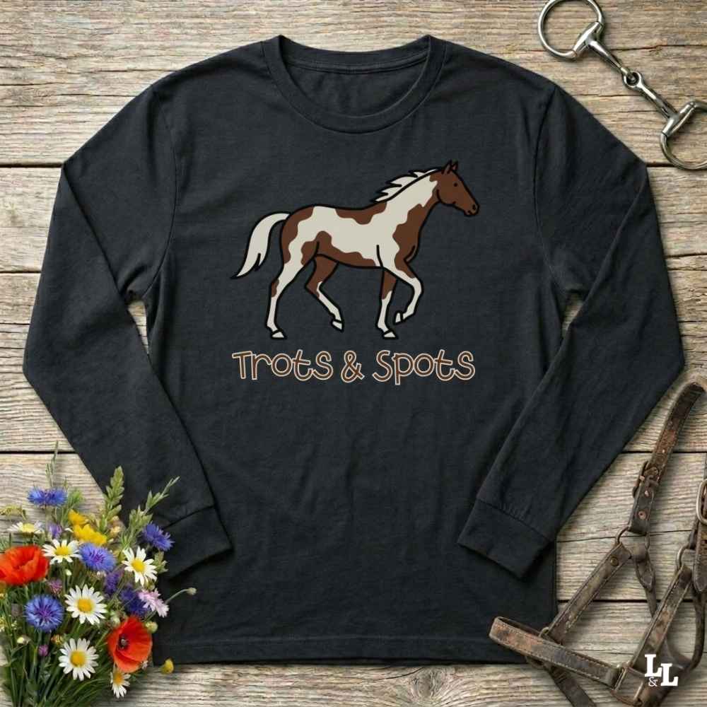 A black long-sleeve t-shirt with a brown and white paint horse graphic and the text 'Trots & Spots' printed on the front, displayed on a rustic wooden background with equestrian accessories and flowers.