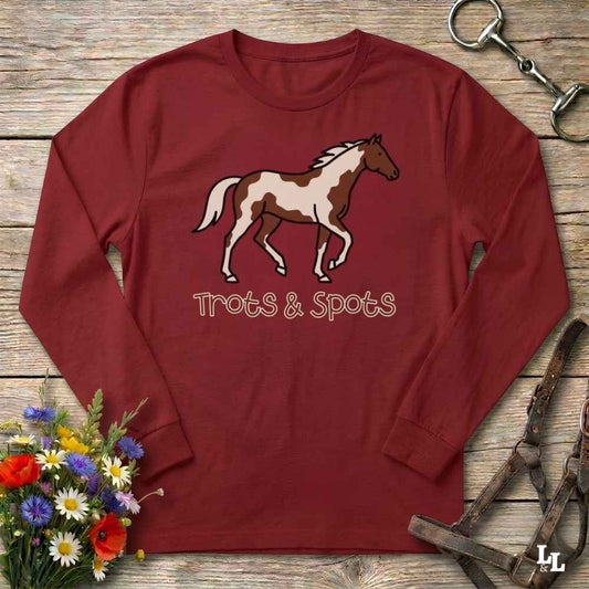 A long-sleeve crewneck t-shirt in maroon red, featuring a graphic of a brown and white paint horse with the text 'Trots & Spots' underneath. The shirt is displayed flat on a rustic wooden background, styled with equestrian tack and a small bouquet of wildflowers.