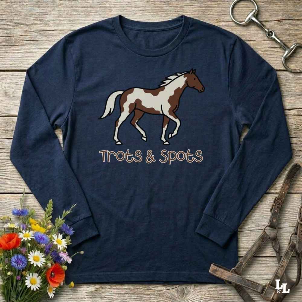 A navy blue long-sleeve t-shirt with a brown and white paint horse graphic and the text 'Trots & Spots' printed on the front, displayed on a rustic wooden background with equestrian accessories and flowers.