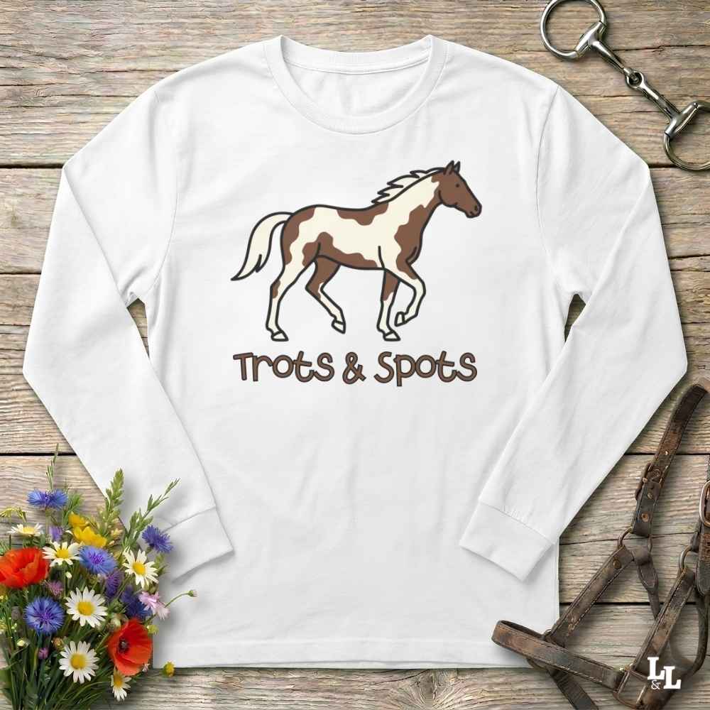 A white long-sleeve t-shirt with a brown and white paint horse graphic and the text 'Trots & Spots' printed on the front, displayed on a rustic wooden background with equestrian accessories and flowers.