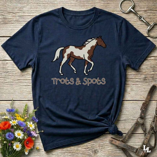 A navy blue graphic t-shirt featuring a brown and white paint horse illustration with the text 'Trots & Spots' underneath, styled in a flat-lay on a rustic wooden background with equestrian accessories and flowers.