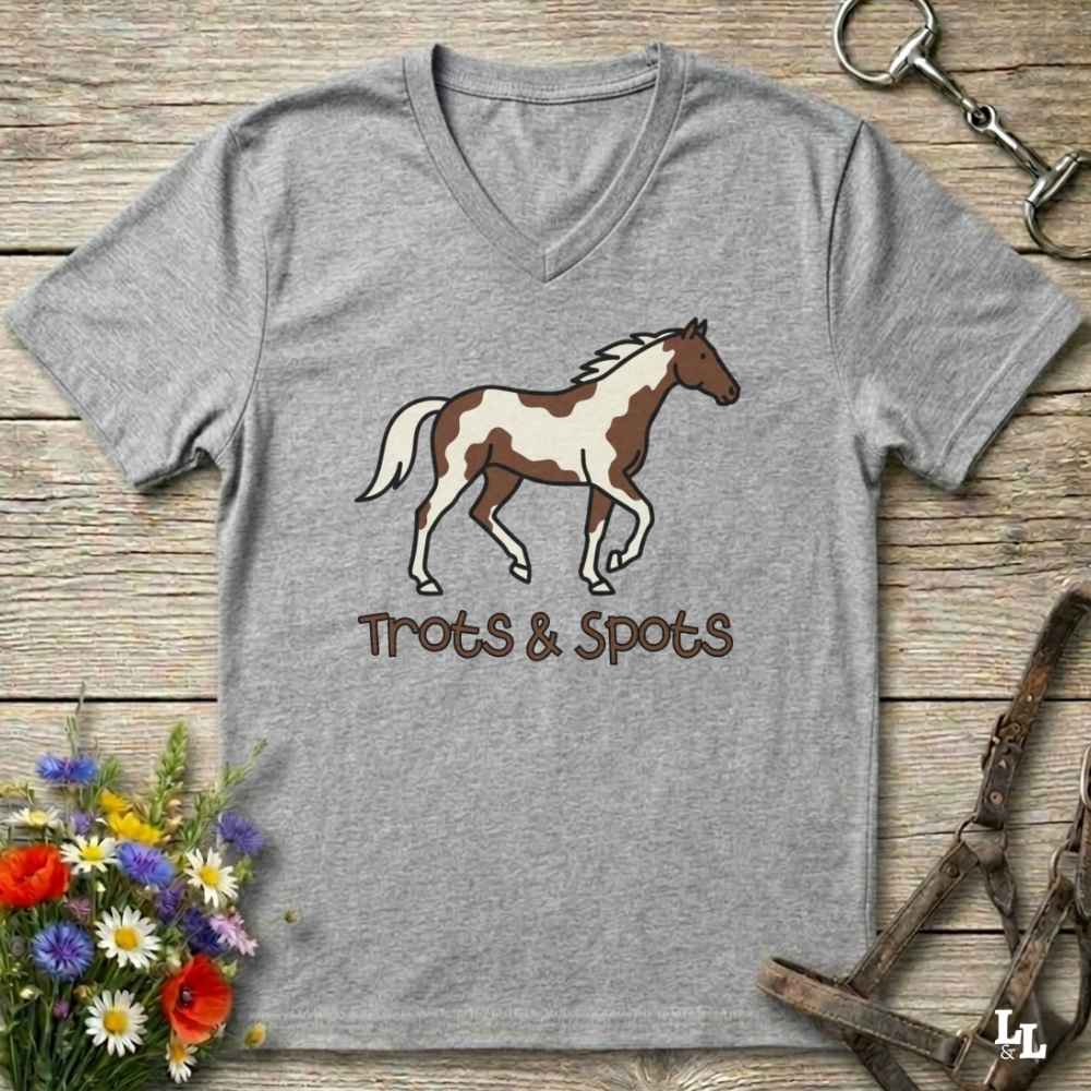 A heather gray V-neck t-shirt featuring a brown and white paint horse graphic with the text 'Trots & Spots' underneath, displayed on a rustic wooden background with equestrian-themed props.
