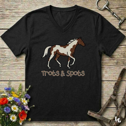 A black V-neck t-shirt featuring a brown and white paint horse graphic with the text 'Trots & Spots' underneath, displayed on a rustic wooden background with equestrian accessories and flowers.