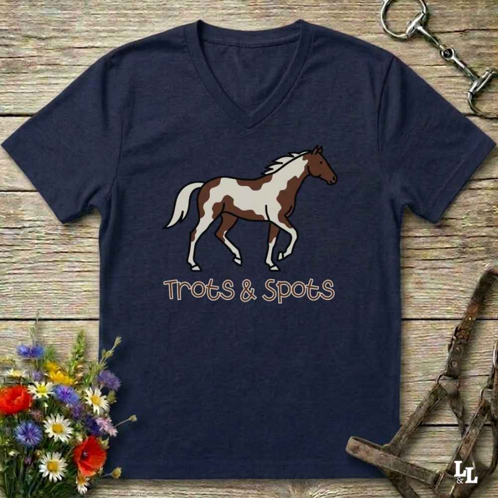 A navy blue V-neck t-shirt featuring a graphic of a brown and white pinto horse with the text 'Trots & Spots' underneath, laid flat on a rustic wooden background with equestrian accessories and flowers.
