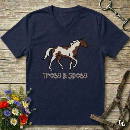 A navy blue V-neck t-shirt featuring a graphic of a brown and white pinto horse with the text 'Trots & Spots' underneath, laid flat on a rustic wooden background with equestrian accessories and flowers.