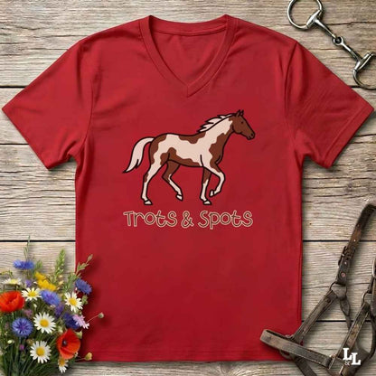 A red V-neck t-shirt laid flat on a rustic wooden surface, featuring a brown and white pinto horse graphic with the text 'Trots & Spots' underneath. The shirt is styled with equestrian accessories (a metal bit in the top right, a leather bridle on the bottom right) and a small bouquet of colorful wildflowers in the bottom left corner.