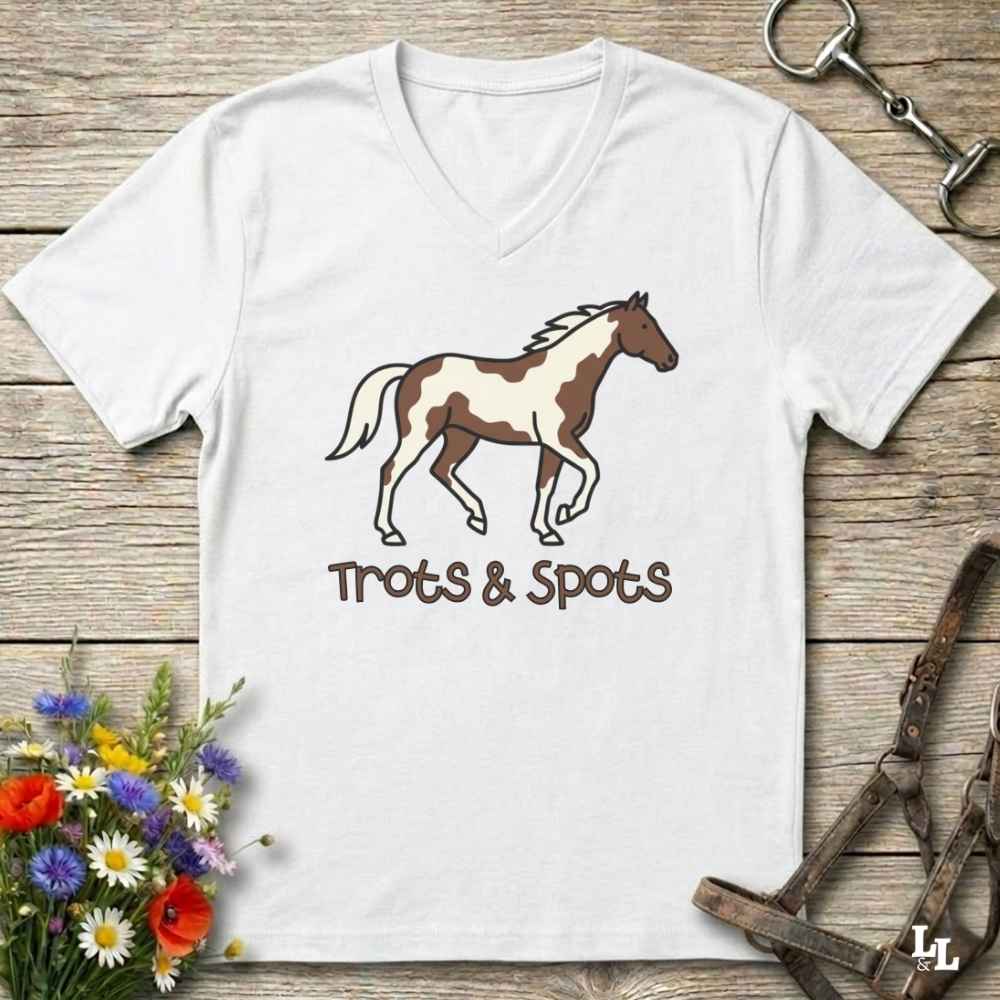 A white V-neck t-shirt laid flat on a rustic wooden surface, featuring a brown and white pinto horse illustration with the text 'Trots & Spots' underneath. The shirt is styled with equestrian accessories (a halter and a horse bit) and a small bouquet of wildflowers.