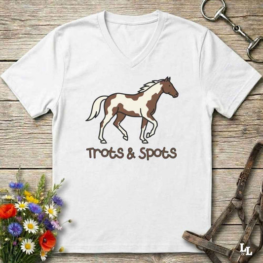 A white V-neck t-shirt laid flat on a rustic wooden surface, featuring a brown and white pinto horse illustration with the text 'Trots & Spots' underneath. The shirt is styled with equestrian accessories (a halter and a horse bit) and a small bouquet of wildflowers.