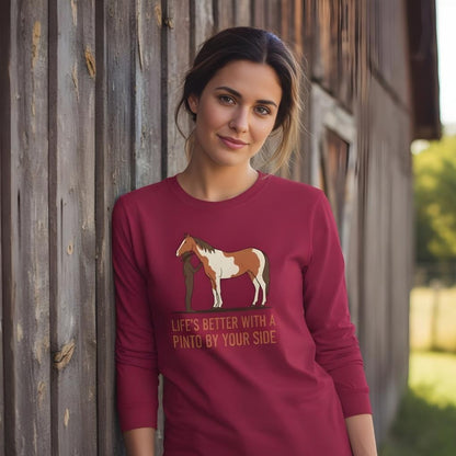 Woman wearing a maroon sweatshirt with a horse graphic and text, standing against a wooden wall. Life's Better with a Pinto Long Sleeve Tee for Pinto Lovers, Trail Riding Tee