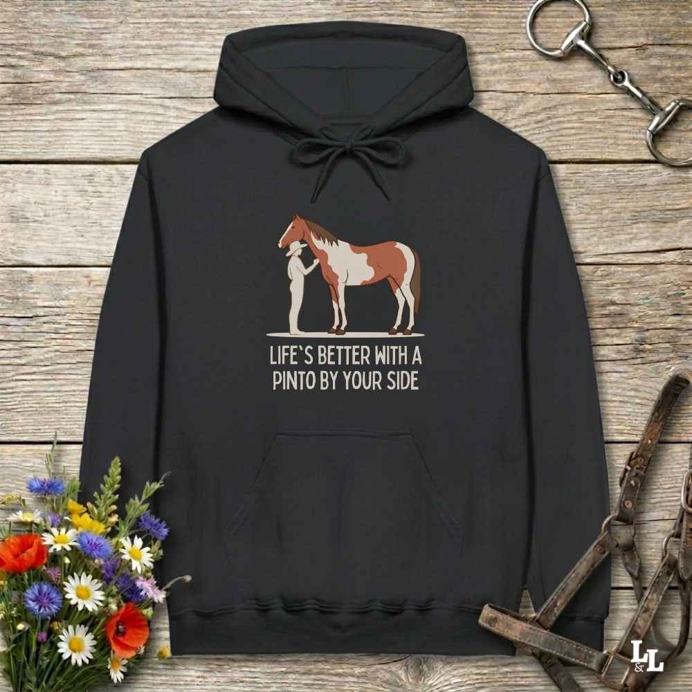 A black pullover hoodie laid flat on a rustic wood surface, featuring a printed graphic of a person standing beside a brown and white Pinto horse, with the text 'LIFE'S BETTER WITH A PINTO BY YOUR SIDE.' Decorative flowers and horse tack are placed around the hoodie.