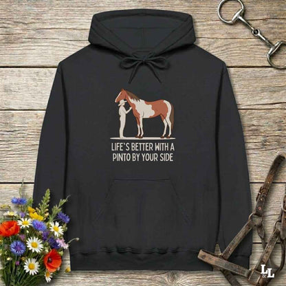 A black pullover hoodie laid flat on a rustic wood surface, featuring a printed graphic of a person standing beside a brown and white Pinto horse, with the text 'LIFE'S BETTER WITH A PINTO BY YOUR SIDE.' Decorative flowers and horse tack are placed around the hoodie.