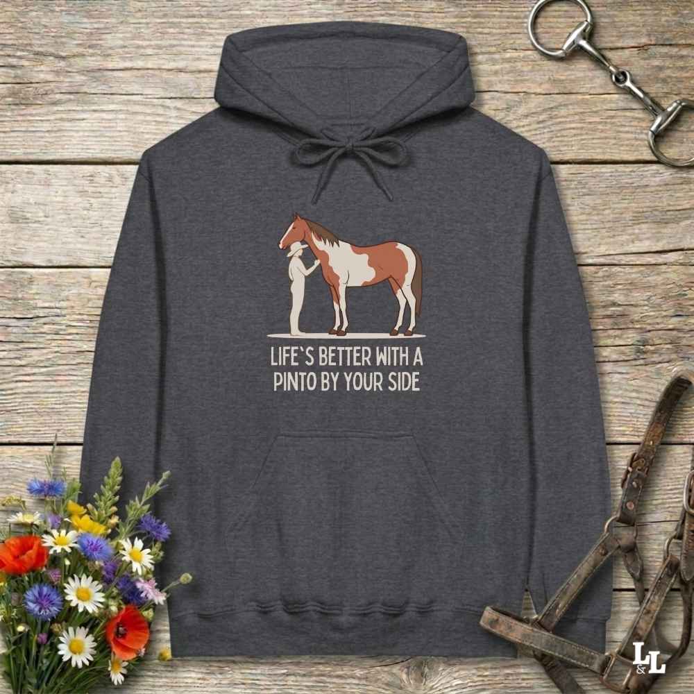 A dark heather gray hoodie laid flat on a rustic wood surface, featuring a graphic of a brown and white pinto horse with a person beside it and the text 'LIFE'S BETTER WITH A PINTO BY YOUR SIDE.' Decorative flowers and horse tack are placed around the hoodie.