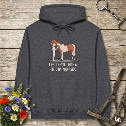 A dark heather gray hoodie laid flat on a rustic wood surface, featuring a graphic of a brown and white pinto horse with a person beside it and the text 'LIFE'S BETTER WITH A PINTO BY YOUR SIDE.' Decorative flowers and horse tack are placed around the hoodie.