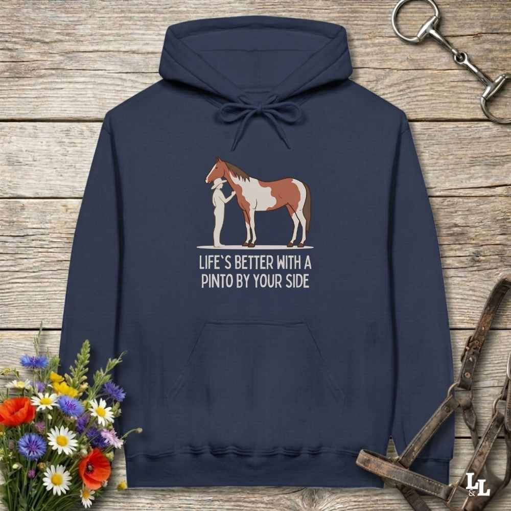 A navy blue hoodie laid flat on a rustic wood background, featuring a printed graphic of a person standing next to a brown and white Pinto horse with the text 'LIFE'S BETTER WITH A PINTO BY YOUR SIDE.' Decorative flowers, horse tack, and a leather strap surround the hoodie.