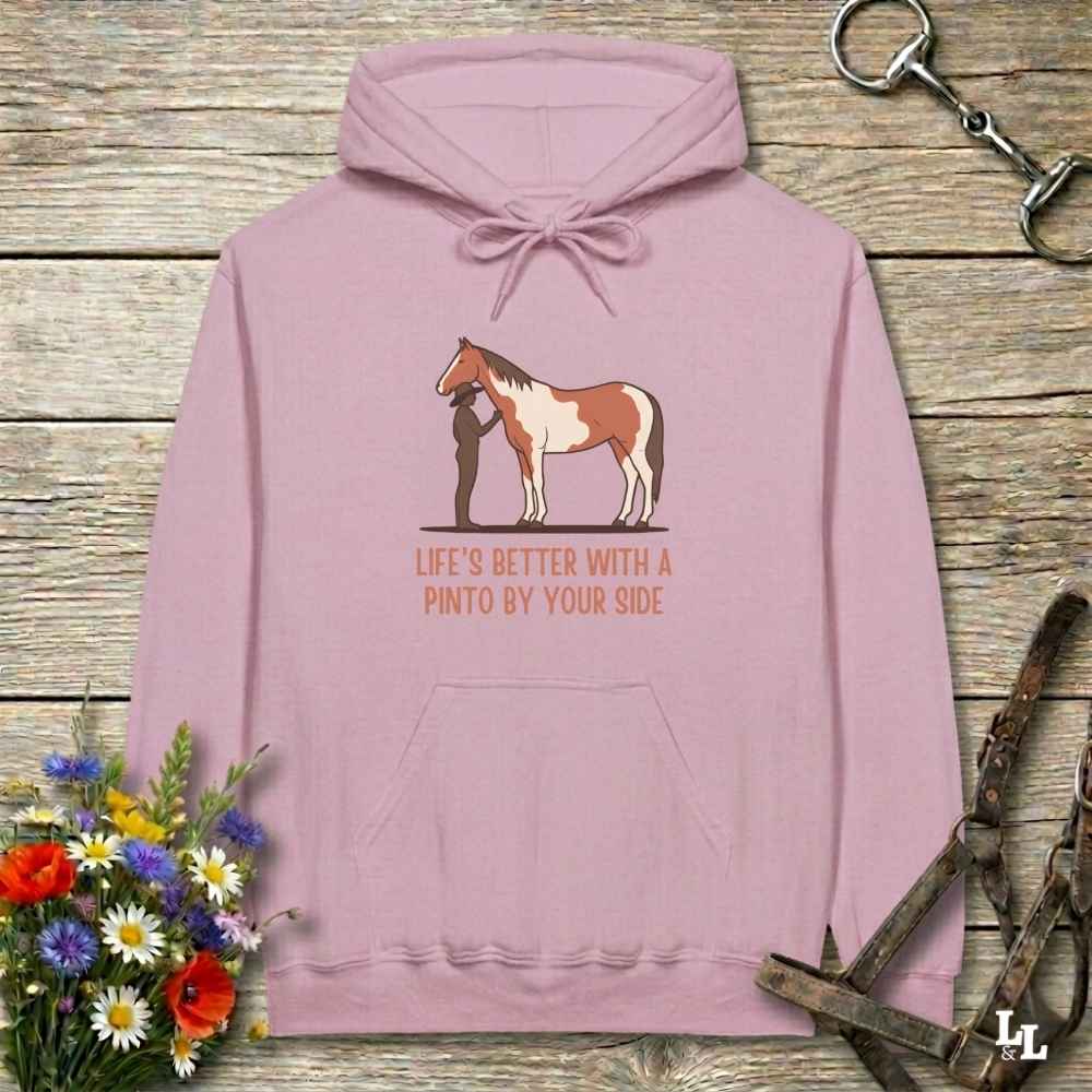 A dusty pink pullover hoodie laid flat on a rustic wooden surface, featuring a graphic of a Pinto horse and a person with the text 'LIFE'S BETTER WITH A PINTO BY YOUR SIDE'. The hoodie is styled with equestrian-themed props including a halter, tack, and a small bouquet of daisies and red flowers.