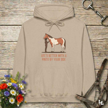 A beige pullover hoodie laid flat on a rustic wood surface, featuring a printed graphic of a person standing beside a paint horse with the text 'LIFE'S BETTER WITH A PINTO BY YOUR SIDE.' Decorative flowers, a horse bit, and a halter are placed around the hoodie.