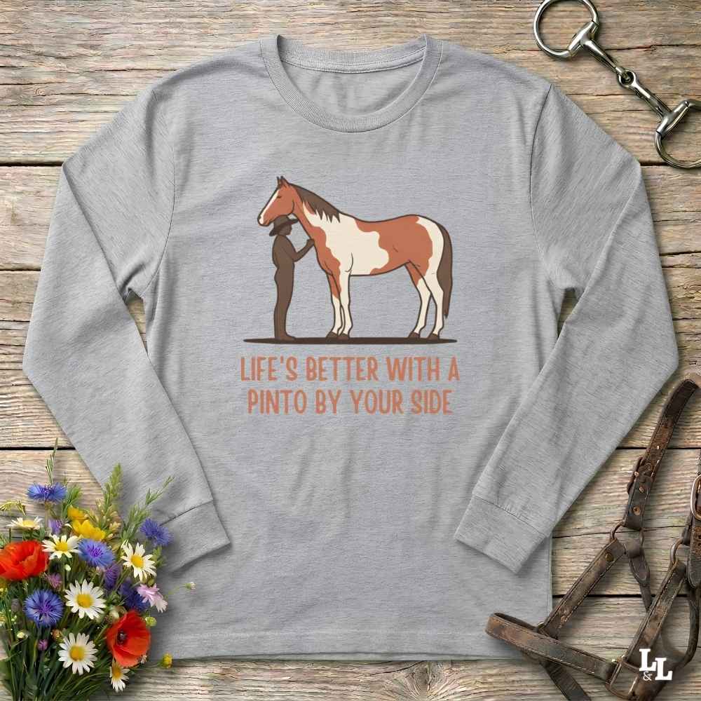 A light gray long-sleeve t-shirt with a graphic of a Pinto horse and a person, accompanied by the text 'LIFE'S BETTER WITH A PINTO BY YOUR SIDE' in orange. The shirt is displayed flat on a rustic wooden background with equestrian accessories and flowers.