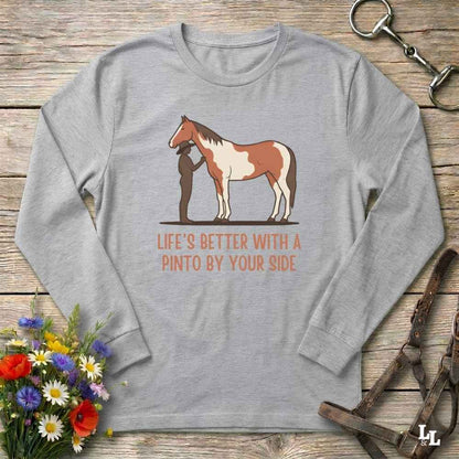 A light gray long-sleeve t-shirt with a graphic of a Pinto horse and a person, accompanied by the text 'LIFE'S BETTER WITH A PINTO BY YOUR SIDE' in orange. The shirt is displayed flat on a rustic wooden background with equestrian accessories and flowers.